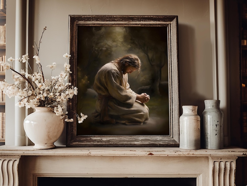 May include: A framed artwork depicts a person kneeling in prayer, set against a backdrop of a forest. The artwork is in a dark wooden frame. A white vase with white flowers and two ceramic jars are on a white mantel.
