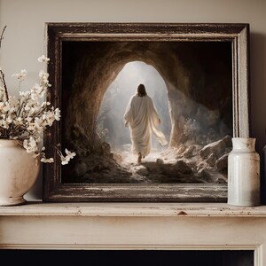 May include: A framed wall art print depicting a figure in a white robe walking toward a bright light at the entrance of a rocky cave. The artwork is displayed on a mantelpiece next to a white vase filled with white flowers and two ceramic vases.