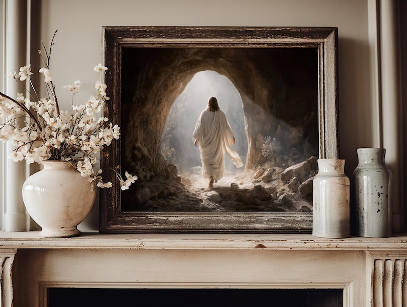 May include: A framed wall art print depicting a figure in a white robe walking toward a bright light at the entrance of a rocky cave. The artwork is displayed on a mantelpiece next to a white vase filled with white flowers and two ceramic vases.