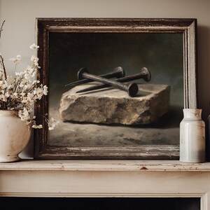 May include: A framed oil painting depicting three dark, weathered iron nails resting on a rough, rectangular stone block. The artwork is displayed on a mantelpiece alongside a white ceramic vase filled with small white flowers and two ceramic bottles.