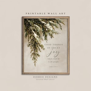 Puede incluir: Un cuadro rectangular enmarcado en madera con una rama de pino nevada que cuelga de la esquina superior izquierda. El texto dice: GOOD TIDINGS OF GREAT joy which shall be to all people. LUKE 2:10-11. El fondo es de un color beige neutro.