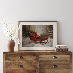 May include: A framed print of a red antique sleigh decorated with evergreen boughs and red bows, set in a snowy forest scene. The artwork is displayed on a wooden dresser with a vase of white flowers.