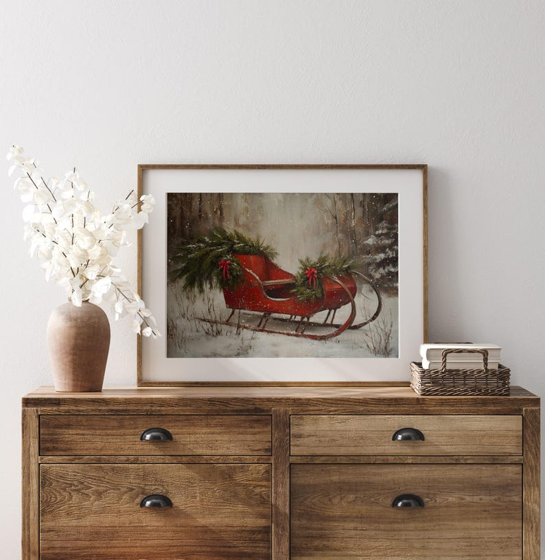 May include: A framed print of a red antique sleigh decorated with evergreen boughs and red bows, set in a snowy forest scene. The artwork is displayed on a wooden dresser with a vase of white flowers.