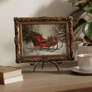 May include: A framed painting of a red sleigh in a snowy forest scene. The painting is in an ornate gold frame and is displayed on a small metal easel. The sleigh is decorated with greenery and red bows, evoking a winter holiday theme.