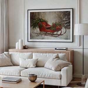 May include: A framed print of a red sleigh decorated with greenery and red bows in a snowy forest scene. The artwork is displayed above a light wood cabinet and a white sofa with beige pillows.