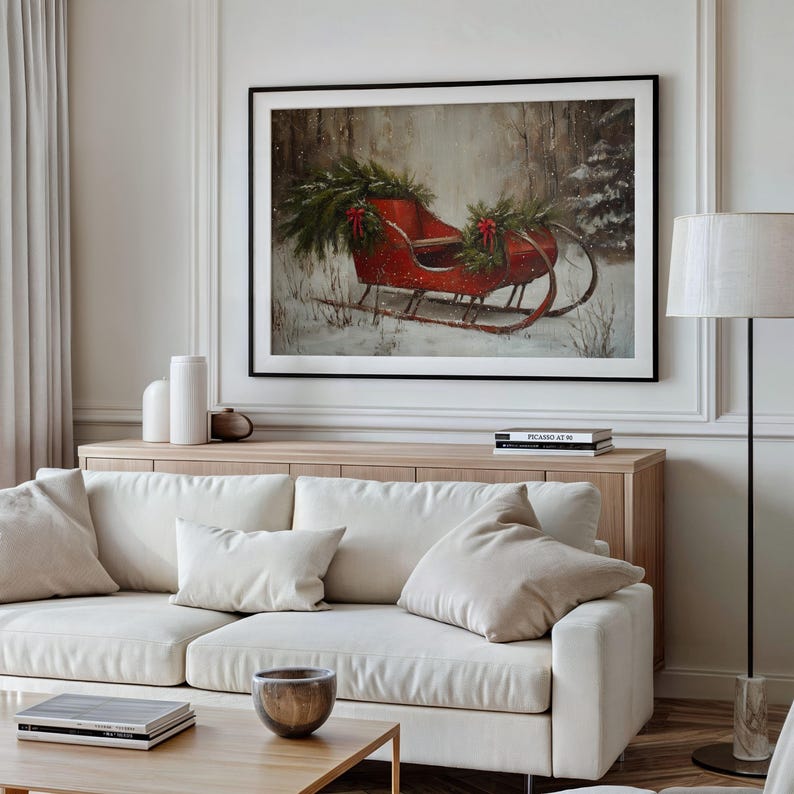 May include: A framed print of a red sleigh decorated with greenery and red bows in a snowy forest scene. The artwork is displayed above a light wood cabinet and a white sofa with beige pillows.