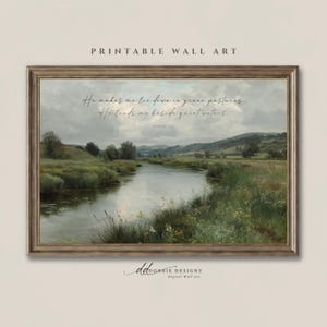 May include: A framed landscape wall art print featuring a river flowing through green pastures under a cloudy sky. The text reads: He makes me lie down in green pastures. He leads me beside quiet waters. Psalm 23:2. The frame is a metallic bronze color.