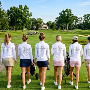 May include: Six women walking on a golf course, wearing white long-sleeved shirts with text and a bird graphic on the back. They are wearing golf skirts and shorts in various colors. Golf clubs and golf carts are visible in the background.