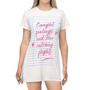 Op de afbeelding: Een wit t-shirtjurkje met een blauw gelijnd notitiepapierpatroon op de voorzijde. De tekst "Caught feelings. and Now catching flights" is gedrukt in een roze cursief lettertype, vergezeld van twee kleine roze hartjes en een roze onderstreping.