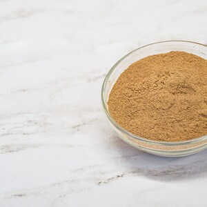 May include: A clear glass bowl filled with brown amla powder sits on a white marble surface. The text "organic way" and "AMLA POWDER" are in the upper left corner. The powder is finely ground and fills the bowl.