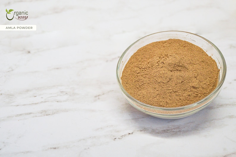 May include: A clear glass bowl filled with brown amla powder sits on a white marble surface. The text "organic way" and "AMLA POWDER" are in the upper left corner. The powder is finely ground and fills the bowl.