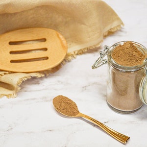 May include: A glass jar filled with brown powder, next to a golden spoon holding a scoop of the same powder. A wooden slotted spoon and a beige cloth are in the background. The text "Organic Way" and "Amla Powder" are visible.