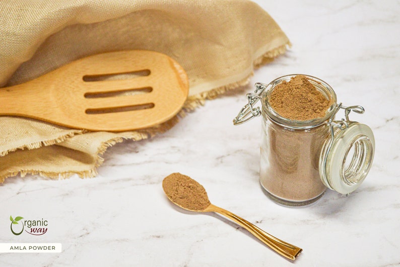 May include: A glass jar filled with brown powder, next to a golden spoon holding a scoop of the same powder. A wooden slotted spoon and a beige cloth are in the background. The text "Organic Way" and "Amla Powder" are visible.