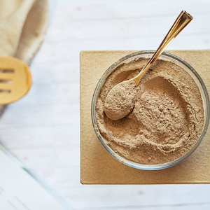 May include: A top-down shot of a clear glass bowl filled with brown amla powder, with a gold spoon resting inside. The bowl sits on a small, square, light brown surface. A wooden spoon and a book are in the background. The text "AMLA POWDER" is visible.