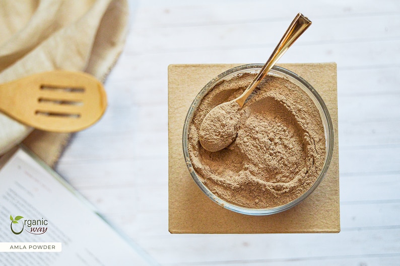 May include: A top-down shot of a clear glass bowl filled with brown amla powder, with a gold spoon resting inside. The bowl sits on a small, square, light brown surface. A wooden spoon and a book are in the background. The text "AMLA POWDER" is visible.