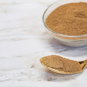May include: A close-up shot of amla powder in a clear glass bowl and on a gold spoon. The powder is a light brown color. The text "AMLA POWDER" is in a white box in the upper left corner.