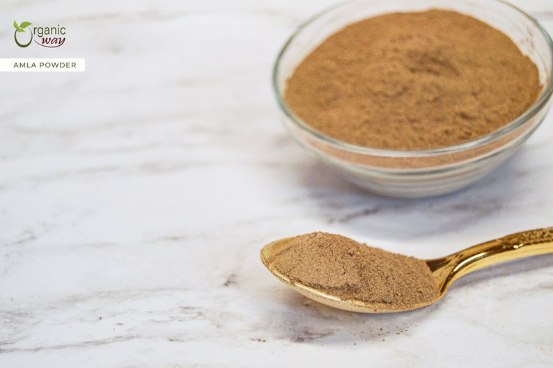 May include: A close-up shot of amla powder in a clear glass bowl and on a gold spoon. The powder is a light brown color. The text "AMLA POWDER" is in a white box in the upper left corner.