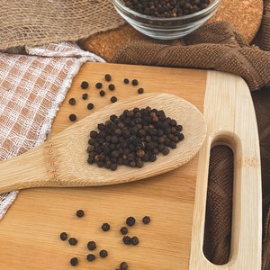 Puede incluir: Una cuchara de madera llena de granos de pimienta negra sobre una tabla de cortar de madera. Tambi&eacute;n hay granos de pimienta esparcidos alrededor de la cuchara. El texto "organic way BLACK PEPPER WHOLE" es visible en la esquina superior izquierda de la imagen.