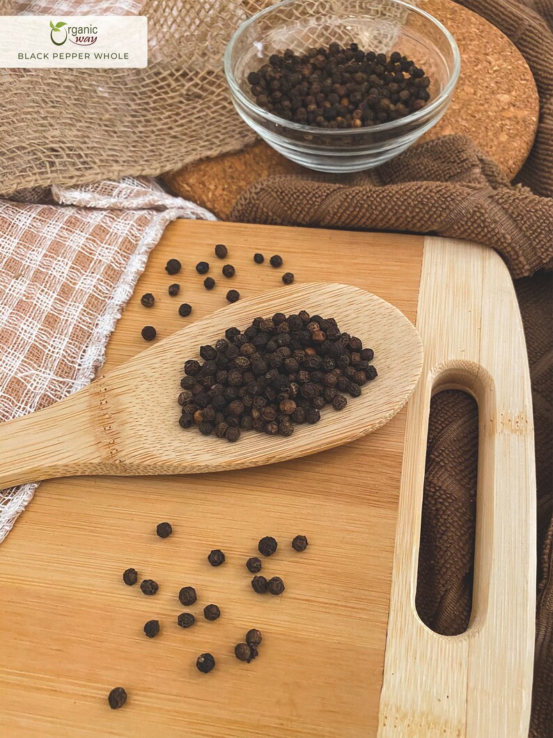 Puede incluir: Una cuchara de madera llena de granos de pimienta negra sobre una tabla de cortar de madera. Tambi&eacute;n hay granos de pimienta esparcidos alrededor de la cuchara. El texto "organic way BLACK PEPPER WHOLE" es visible en la esquina superior izquierda de la imagen.