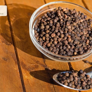 Puede incluir: Un bol de vidrio y una cuchara de metal llenos de granos de pimienta negra enteros. Los granos de pimienta son de color marr&oacute;n oscuro y est&aacute;n esparcidos sobre una superficie de madera.