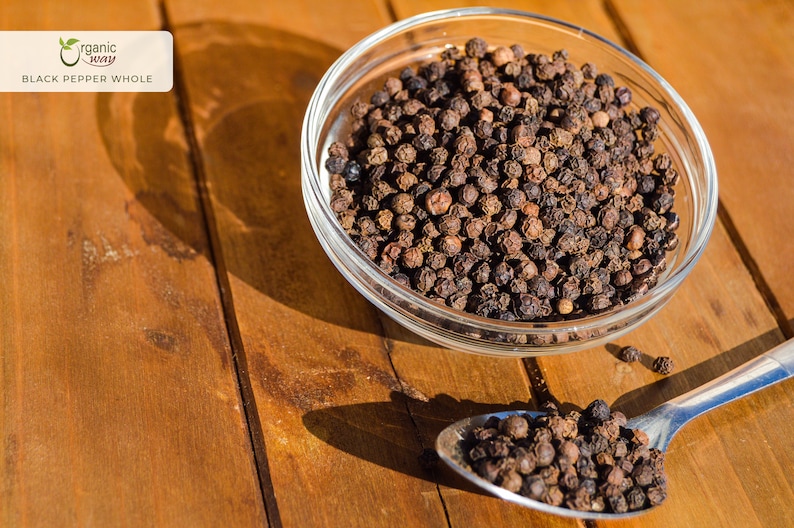 Puede incluir: Un bol de vidrio y una cuchara de metal llenos de granos de pimienta negra enteros. Los granos de pimienta son de color marr&oacute;n oscuro y est&aacute;n esparcidos sobre una superficie de madera.