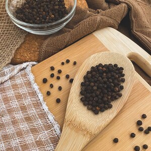 Puede incluir: Una cuchara de madera llena de granos de pimienta negra, con granos de pimienta adicionales esparcidos sobre una superficie de madera. El texto "organic way BLACK PEPPER WHOLE" es visible en la esquina superior derecha de la imagen.