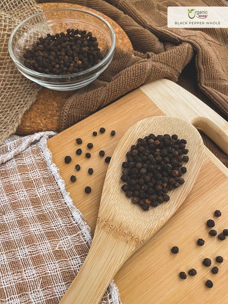 Puede incluir: Una cuchara de madera llena de granos de pimienta negra, con granos de pimienta adicionales esparcidos sobre una superficie de madera. El texto "organic way BLACK PEPPER WHOLE" es visible en la esquina superior derecha de la imagen.