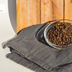 Puede incluir: Un bol de vidrio lleno de granos de pimienta negra enteros, sentado sobre una tela rayada gris y verde. El bol est&aacute; delante de un fondo de madera.