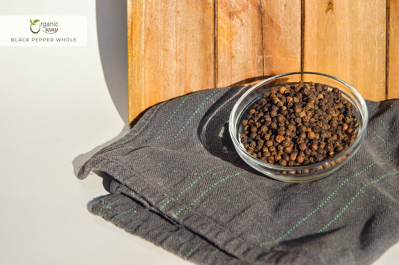 Puede incluir: Un bol de vidrio lleno de granos de pimienta negra enteros, sentado sobre una tela rayada gris y verde. El bol est&aacute; delante de un fondo de madera.