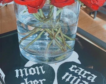 May include: Two black heart-shaped coasters with white lettering. The coaster on the left says "mon cher" and the coaster on the right says "cara mia".