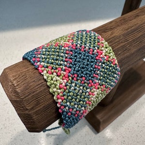May include: A colorful woven bracelet with pink, green, and blue threads. The bracelet is displayed on a wooden stand.