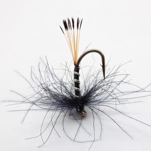 May include: A close-up of a fly fishing lure. The hook is silver with a black body wrapped in silver wire. The lure has black feathers and a cluster of tan and black fibers extending upwards.
