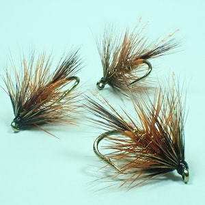Puede incluir: Tres señuelos de pesca con mosca marrones y naranjas con anzuelos dorados.