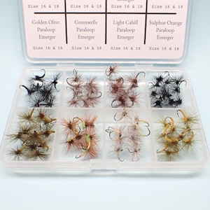 May include: A clear plastic storage box containing eight compartments filled with various fishing flies. The lid features printed text labels for Golden Olive, Greenwells, Light Cahill, and Sulphur Orange Paraloop Emerger flies in sizes 16 & 18.