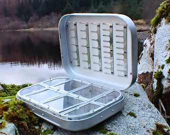 Puede incluir: Caja de aparejos de pesca plateada abierta, que revela un interior blanco y compartimentos transparentes. La caja es rectangular con bordes redondeados. Descansa sobre una roca cubierta de musgo, con un lago y árboles al fondo.