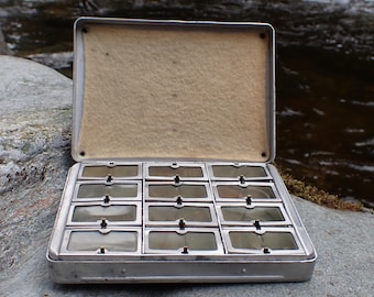 Puede incluir: Un estuche metálico vintage abierto, que revela numerosos compartimentos rectangulares pequeños. El estuche es de color plateado con un forro interior beige. Cada compartimento tiene una cubierta transparente. El estuche descansa sobre una roca gris, con un fondo de agua borroso.