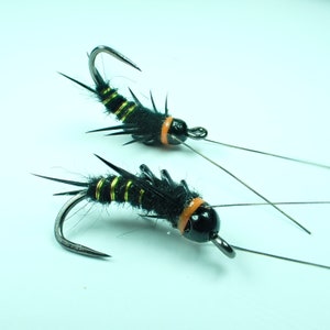 Può includere: Due esche da pesca a mosca nere e gialle con accenti arancioni e ami neri.