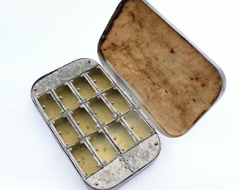 Puede incluir: Una caja de pastillas antigua de metal plateado abierta, con tapa con bisagras. El interior está dividido en doce compartimentos rectangulares con cubiertas transparentes. El interior de la tapa está forrado con un material beige.