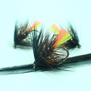 Puede incluir: Tres señuelos de pesca con mosca negros y marrones con plumas naranja, amarillas y verdes.