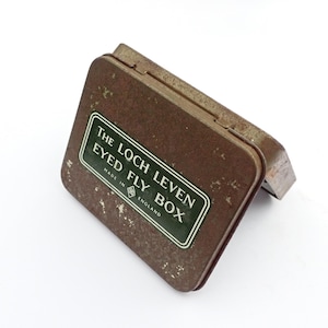 May include: A vintage, rectangular, brown metal fishing fly box with a hinged lid. The box features a green and white label that reads "THE LOCH LEVEN EYED FLY BOX MADE IN ENGLAND". The box has a worn, rustic appearance.