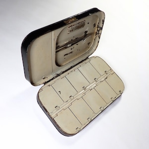 May include: An open, vintage metal paint box. The exterior is black, and the interior is cream-coloured. The rectangular box has rounded edges and compartments for paint. The lid is open, revealing additional storage and a mechanism.