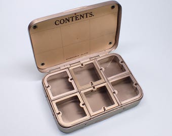Puede incluir: Contenedor metálico vintage abierto con seis compartimentos. La tapa está abierta, revelando la palabra "CONTENTS" y un patrón de cuadrícula. El contenedor es de color dorado apagado, con divisores de plástico transparente.