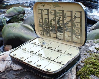 Puede incluir: Una caja de aparejos de pesca con mosca vintage abierta, que revela compartimentos. La caja tiene un borde negro y un interior crema. La tapa tiene múltiples ranuras para guardar moscas. La caja descansa sobre una roca musgosa cerca de un arroyo.