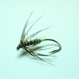 Pode incluir: Uma isca de pesca com um anzol de prata metálica, um corpo verde-oliva escuro envolto em fio dourado e um colarinho verde neon brilhante. A cabeça é escura, adornada com penas longas e finas com manchas pretas e castanhas para fora.