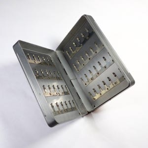 May include: A silver metal fly fishing box with multiple rows of slots for holding flies. The box is open and the slots are visible.