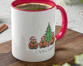 Può includere: Tazza da caffè in ceramica bianca con bordo e manico rossi. La tazza presenta un design natalizio con tre capibara che indossano cappelli da Babbo Natale e tengono dei regali. Il testo "Capy-Christmas" è stampato sulla tazza.