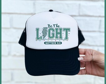 May include: A white and black trucker hat with a green and white design that says "Be the Light" with a lightning bolt and Christmas lights. The text "Matthew 5:14" is below the design.