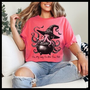 May include: A pink t-shirt with a graphic of an octopus wearing a witch's hat and stirring a cauldron. The text "On My Way To Stir The Pot" is printed below the graphic.