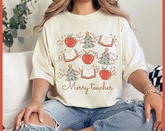 May include: Cream-colored t-shirt with a festive design featuring red apples, green Christmas trees, and open books. The words "Merry teacher" are written in a cursive font. The shirt is worn with blue jeans.