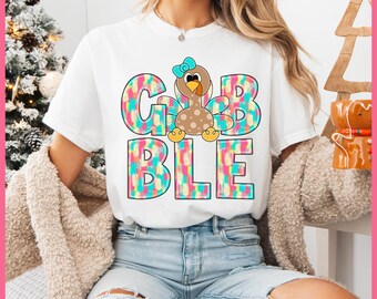 May include: White t-shirt featuring the word "GOBBLE" in colorful, patterned letters, with a cartoon turkey in the center. The turkey has a blue bow and is brown with polka dots. The shirt is worn by a person in blue jeans.